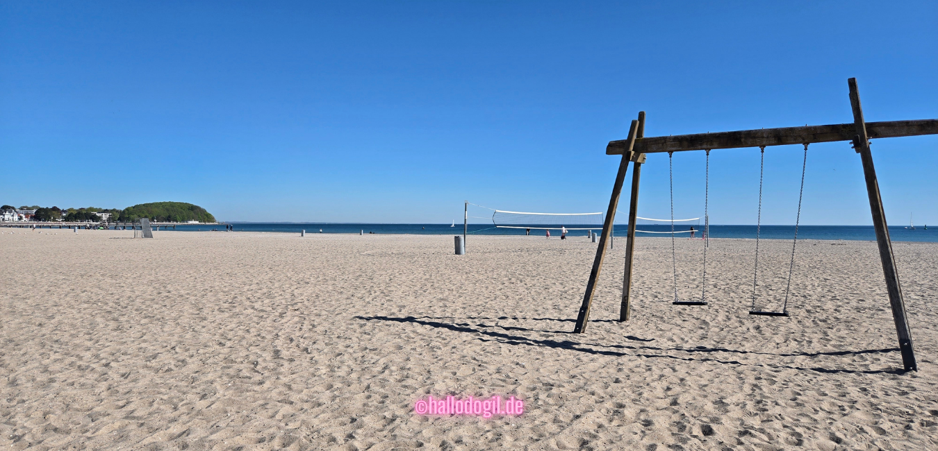 Weiter, breiter und leerer Strand von Travemünde mit Schaukeln im Vordergrund bei schönem blauen Himmel.